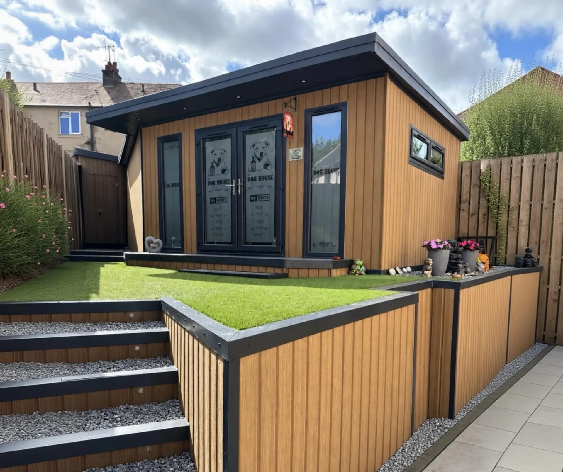 Timber-clad garden room with dog-house themed double doors, artificial grass deck and gravel steps, built as an outdoor entertaining space