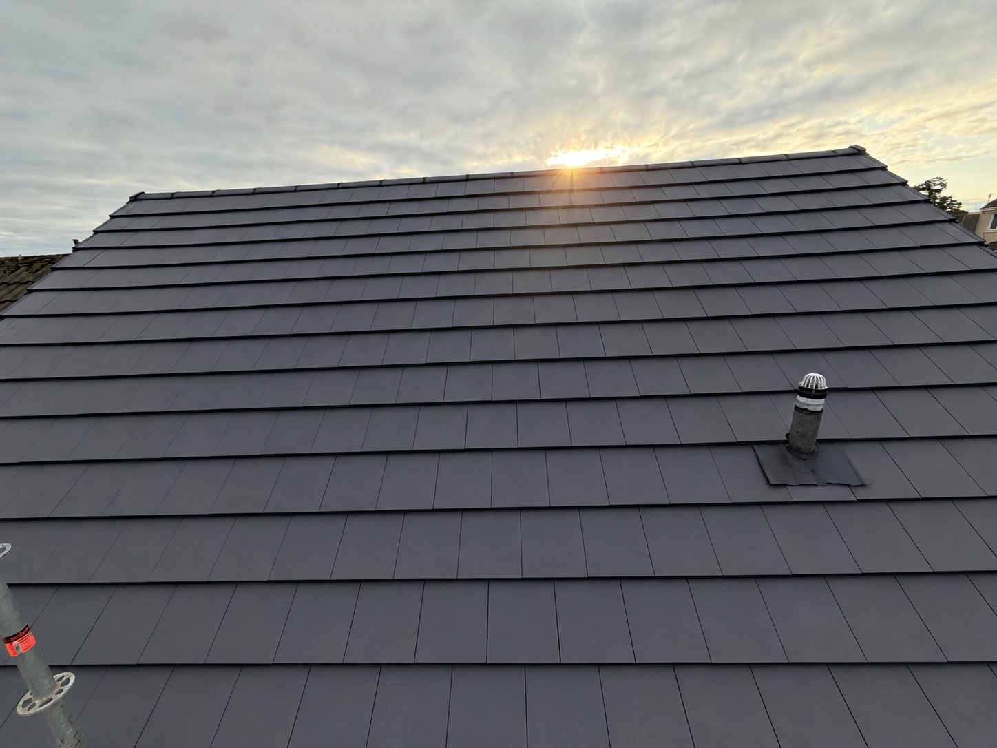 Completed dark grey tiled roof at sunset, with a vent pipe and clean ridge line visible against a cloudy sky